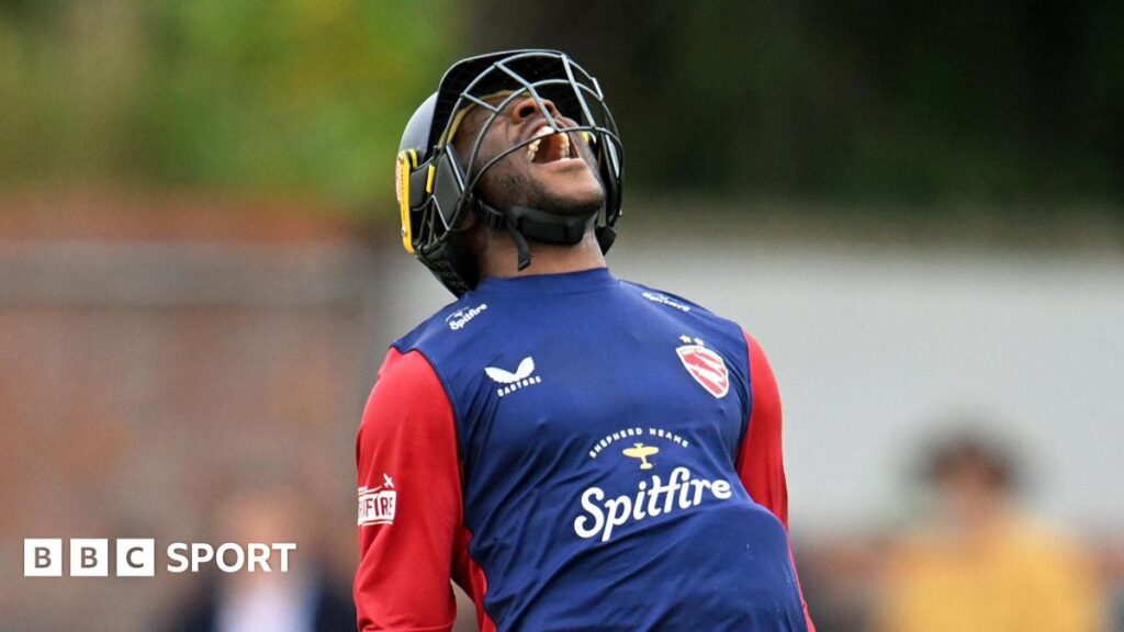 A mid shot of Daniel Bell-Drummond. He is wearing a protective cricket helmet, and a blue shirt with red sleeves. He is roaring with joy after scoring for Kent.