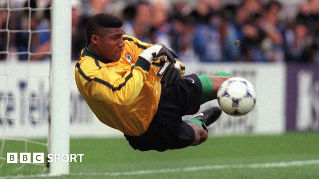 Peter Rufai, wearing a yellow goalkeeping top, dives to his right to save a shot during a football match