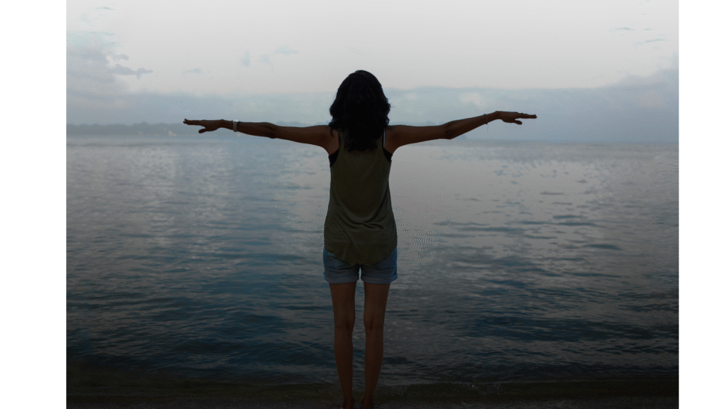 woman standing with outstretched arms along the beach looking at the water and contemplating the weekly horoscope