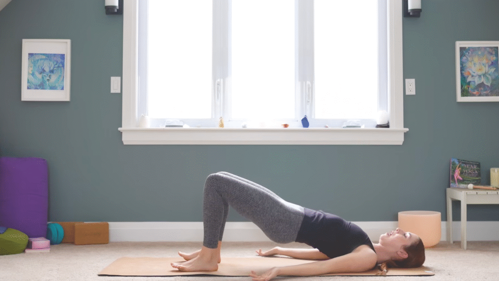 woman in yoga clothes in bridge pose with both feet on mat, knees bent and hips raised toward ceiling on yoga mat in home
