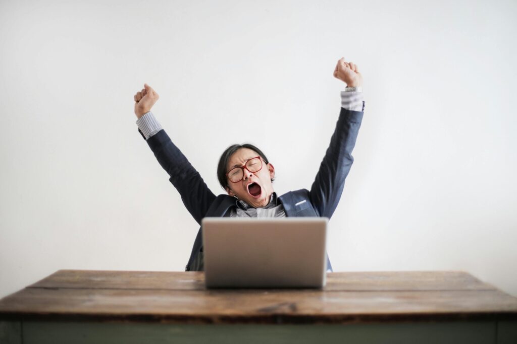 Man practicing desk yoga, sitting at desk with laptop in front of him. His arms are outstretched over his head and he is yawning.