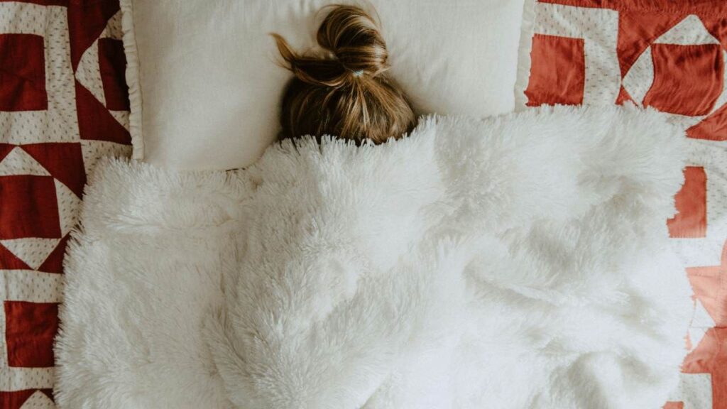 7 mistakes you’re making when trying to keep your bedroom warm in winter Someone with a bun in their hair hiding underneath a white fluffy blanket